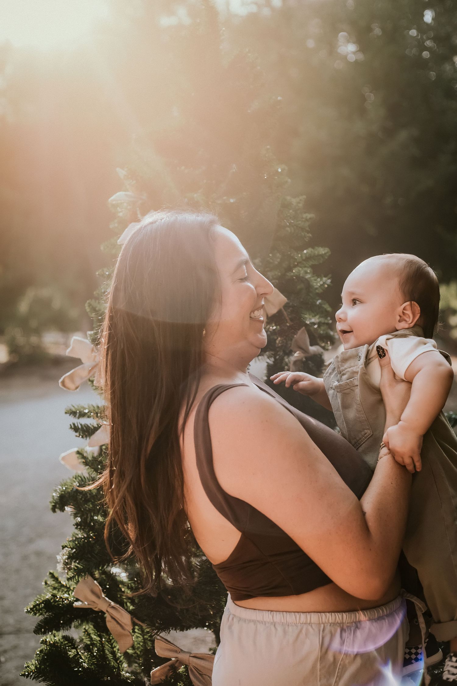 A smiling mother holds her baby outdoors near a Christmas tree with golden sunlight glowing behind them.