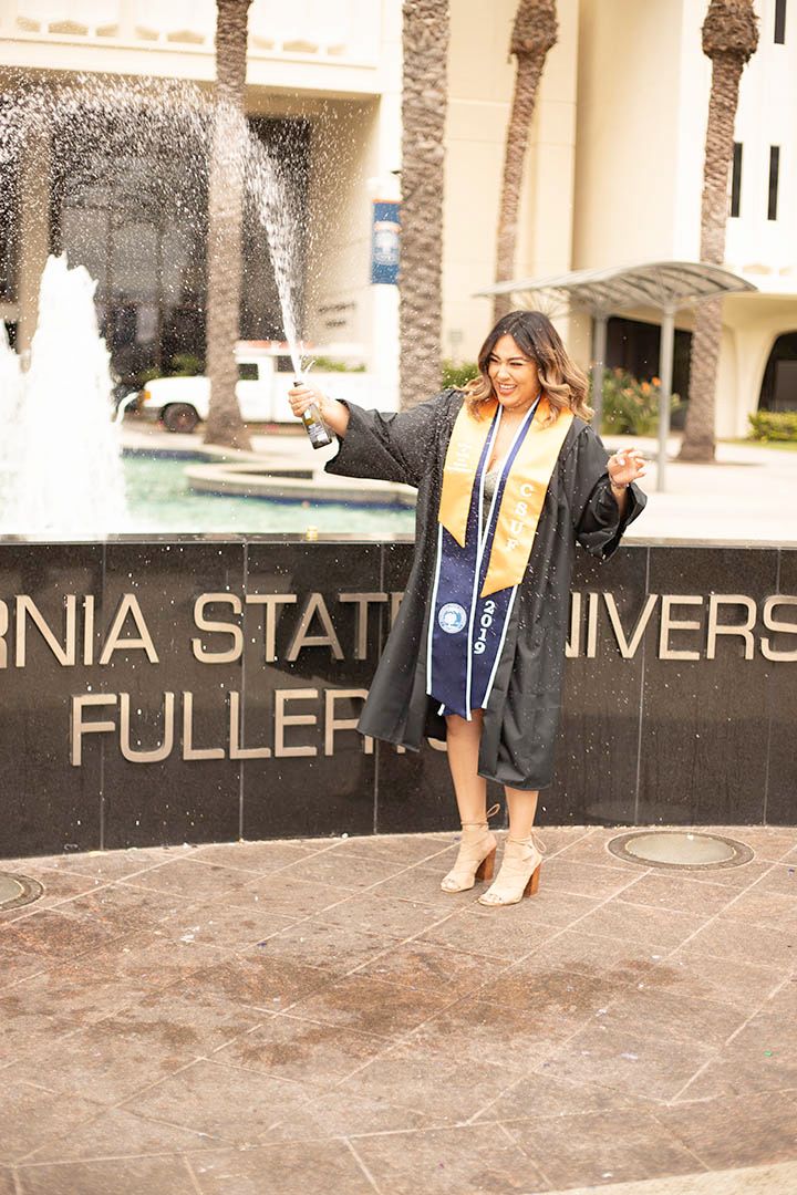 a graduate in her gown laughing while popping champagne in front of a CSUF sign. There is a fountain in the background