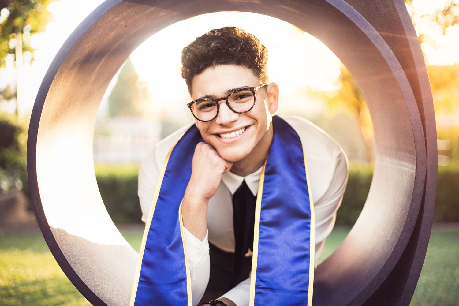 Smiling graduate in blue stole poses playfully through large circular sculpture outdoors.