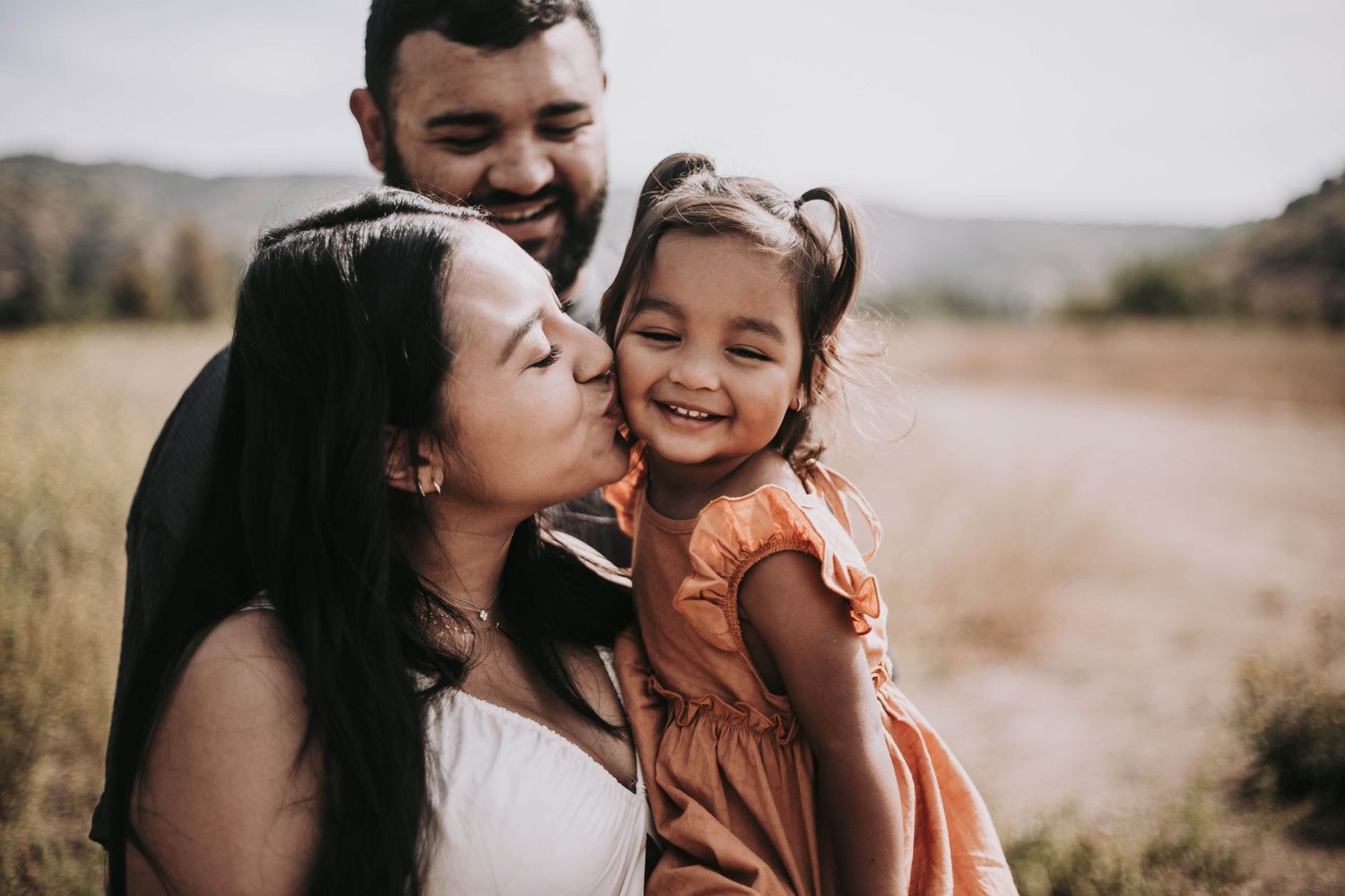 Tender family scene in a rustic field where a mom holds and kisses her toddler daughter who is smiling. Dad is looking at them and smiling