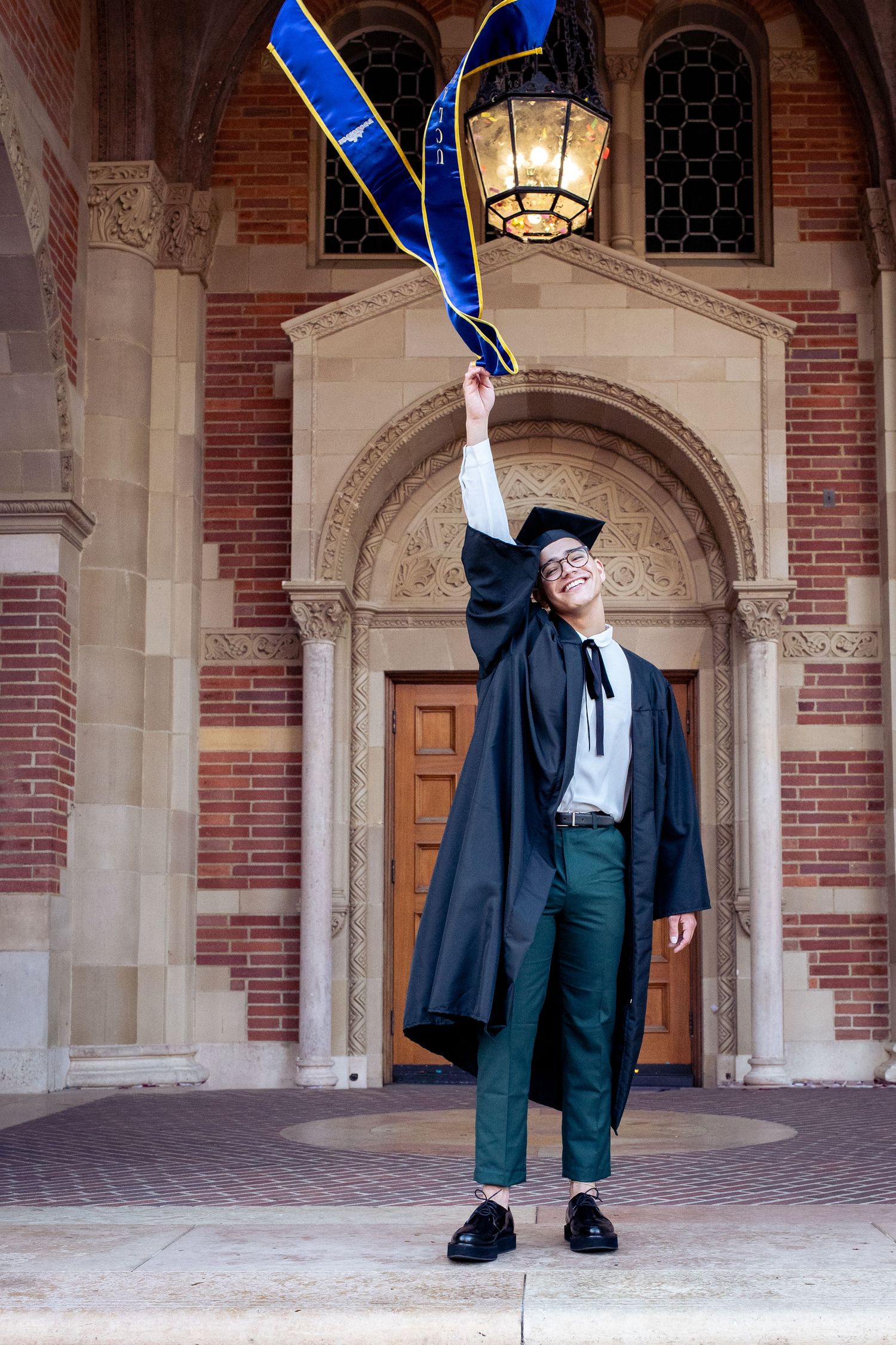 Joyful UCLA graduate in black cap and gown tossing blue stole outside Kerckhoff Hall.