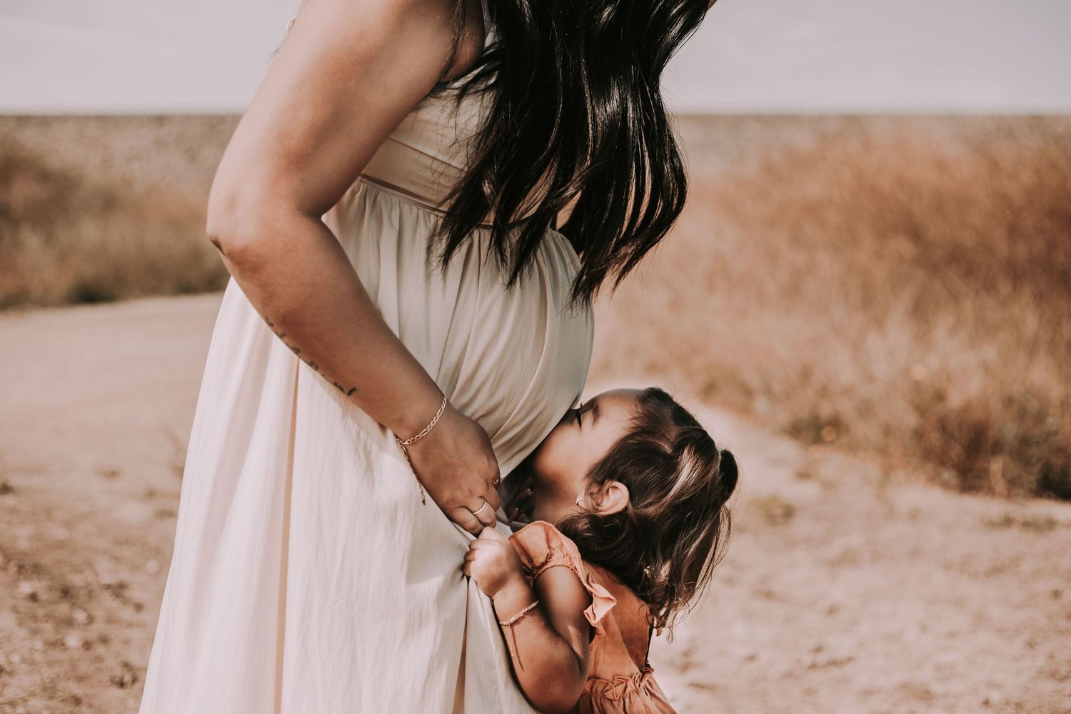 pregnant mother in a flowing dress looks down at her toddler daughter kissing her baby bump in a rustic field during a maternity session