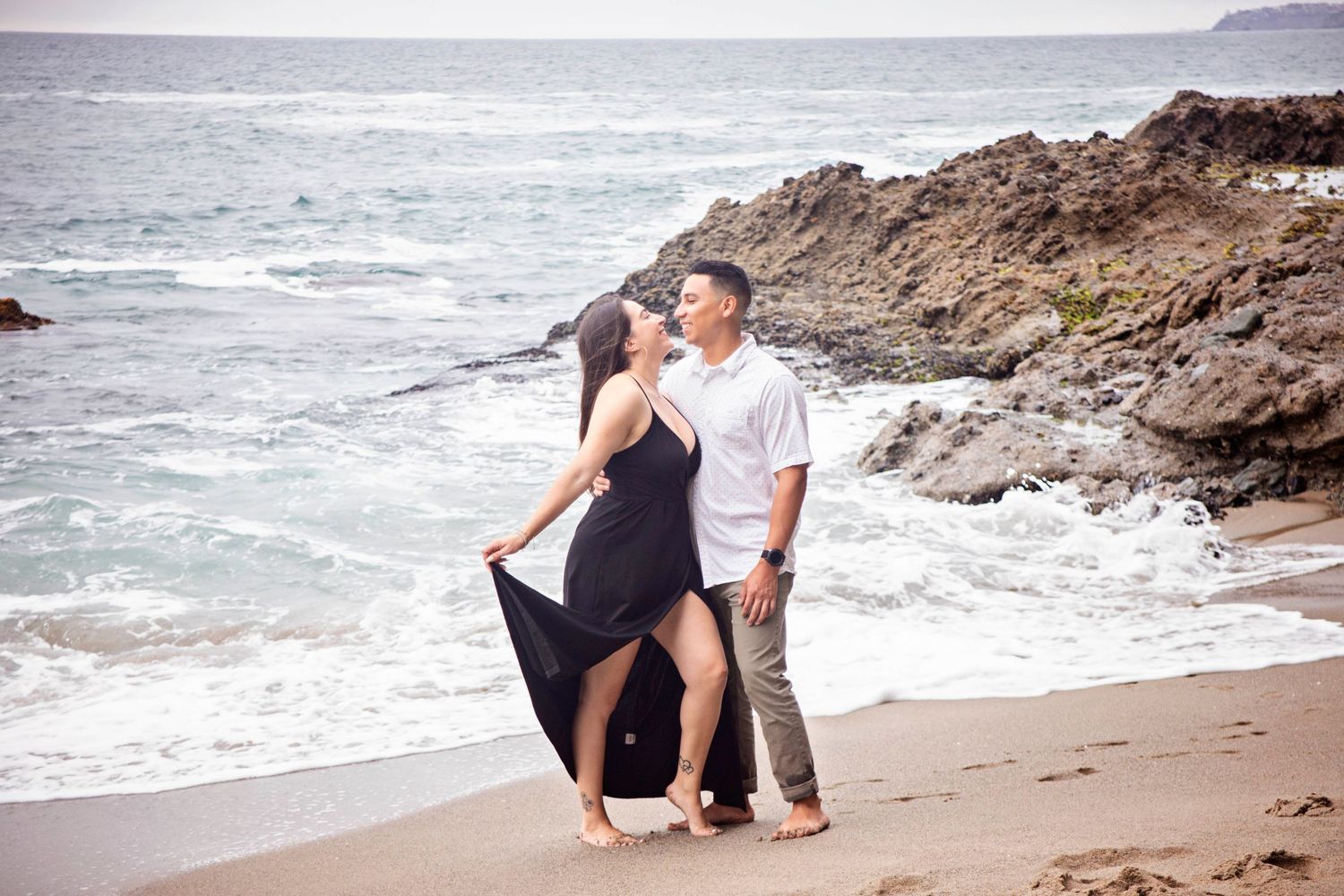 Couple embracing on a sandy beach with ocean waves and rocky cliffs in Laguna Beach, California, during a romantic engagement session