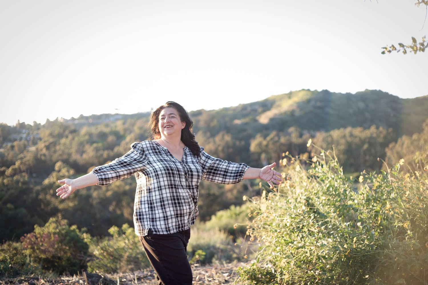 woman is standing in a grassy field at sunset with hills behind her as she twirls and smiles while looking off into the distance off camera
