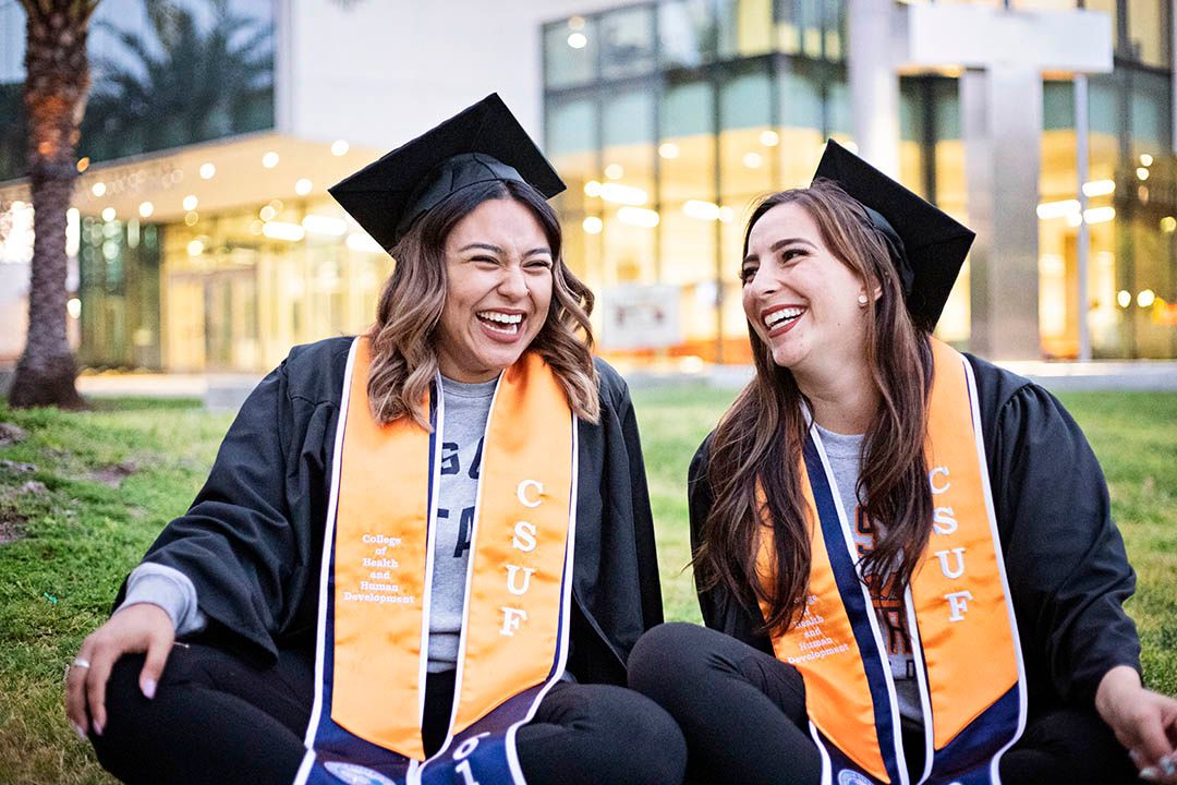 2 CSUF graduates in full graduation attire sitting on a grassy lawn laughing and smiling together at dusk; lit up building in background