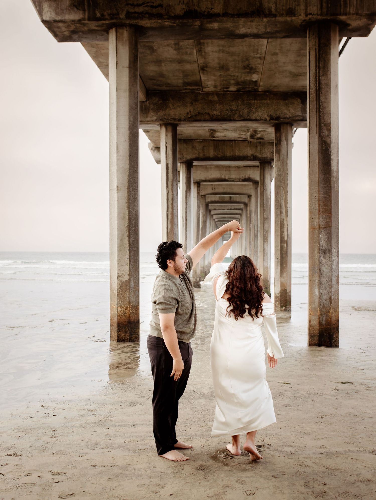 engaged couple dancing under Scripps Pier at the beach. Her back is to the camera as she spins while holding her partner's hand.