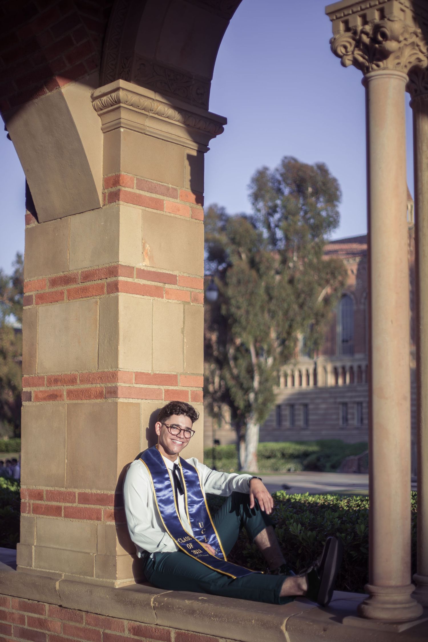 Smiling UCLA graduate sitting against stone column under campus archway in sunlight.