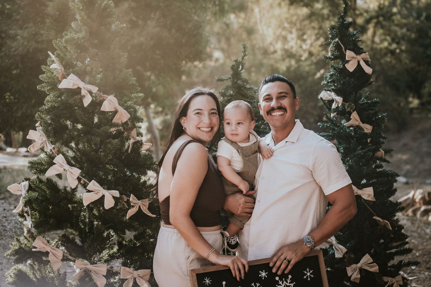 Family of three posing outdoors near decorated Christmas trees, parents smiling and holding their baby in a holiday portrait.