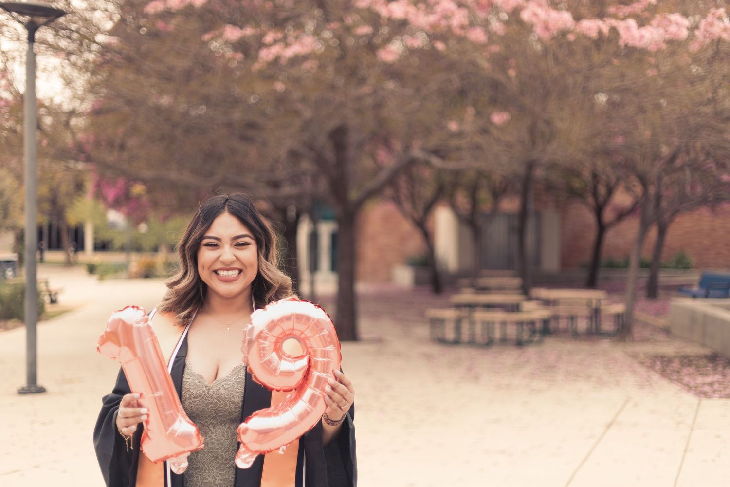 CSUF graduate wearing gown and sash as she holds pink number balloons that show her graduation year (19). She is smiling at the camera