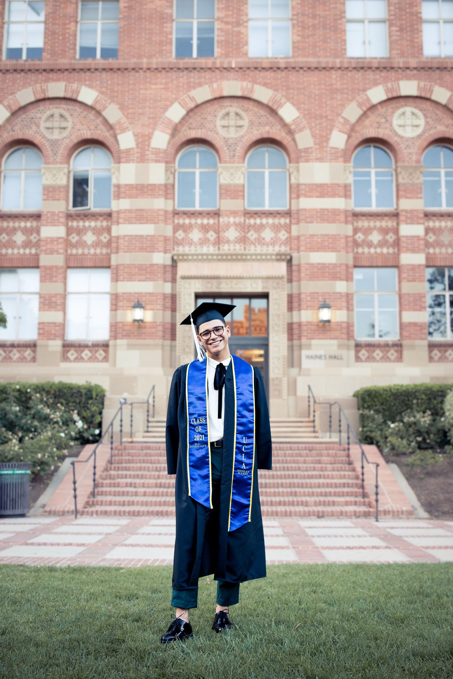 Smiling graduate in cap and gown stands on lawn before UCLA brick building.