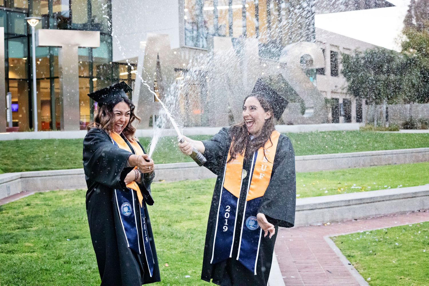 Two graduates in caps and gowns celebrate with champagne spray near campus fountain.