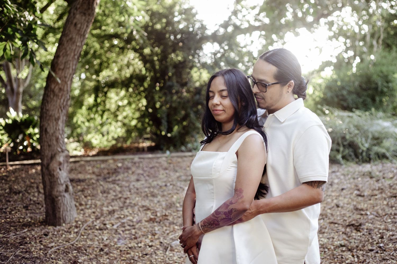 Couple standing in a wooded area; man embraces the woman from behind. Both of them have their eyes closed as they soak in the tender moment