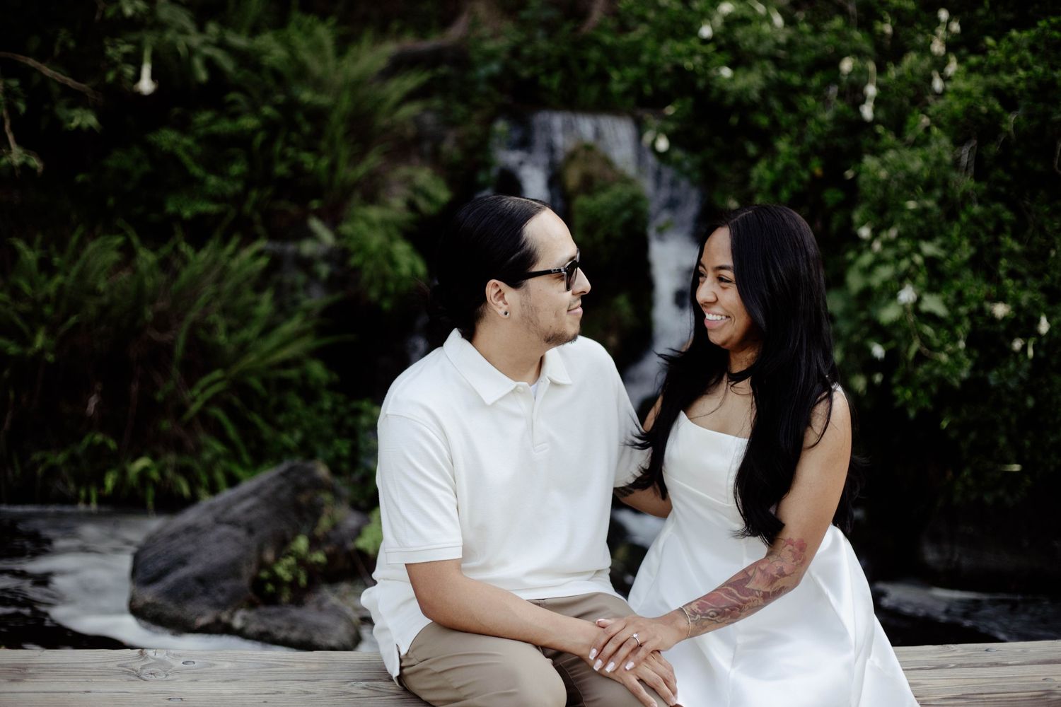 couple sitting on a ledge, holding hands and smiling at each other during an engagement session, with a waterfall in the background