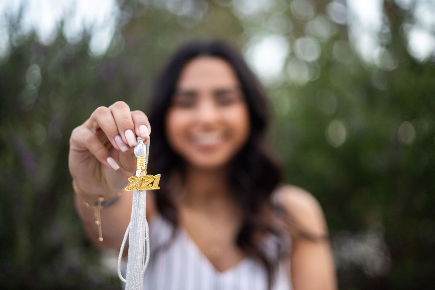college graduate with long hair smiles while holding her tassel for the camera. She is slightly blurry and the tassel is in sharp focus