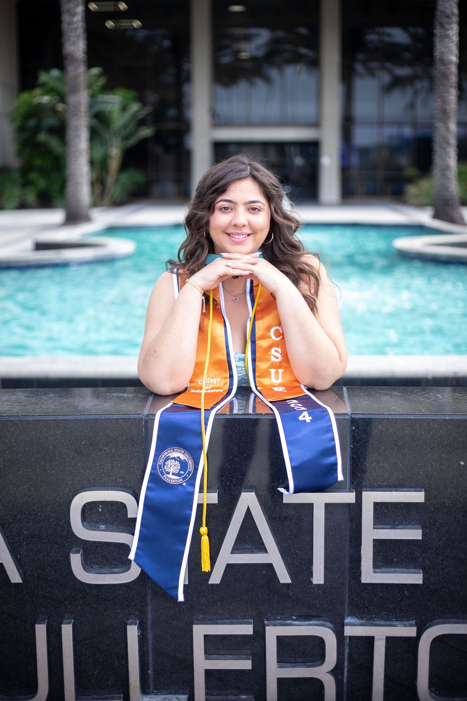 CSUF graduate with her elbows leaning on a CSUF sign during a graduate session. She is smiling at the camera, her hands under her chin