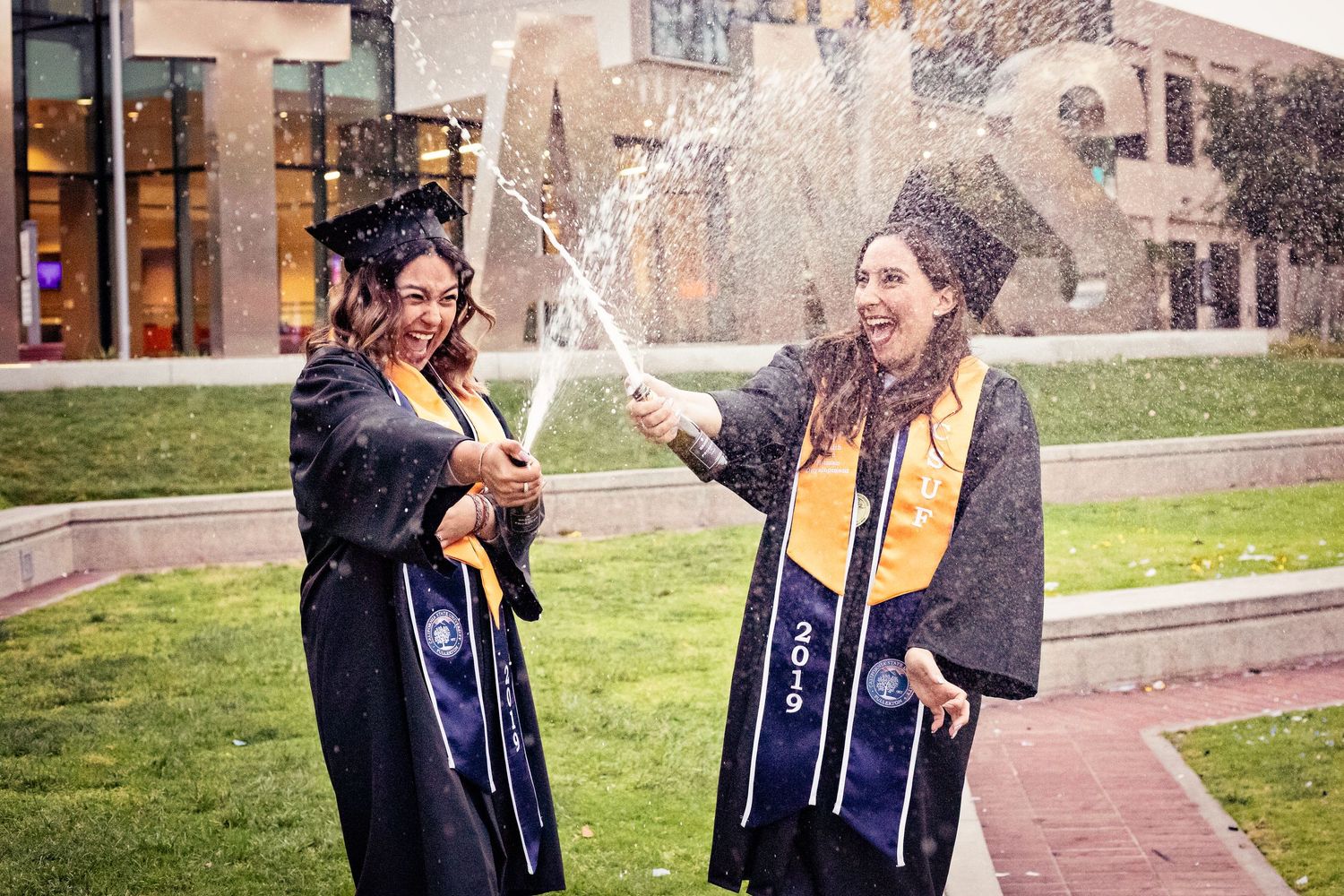 2 CSUF graduates in full graduation attire stand as they pop champagne and laugh on a grassy field with the Titans sign behind them