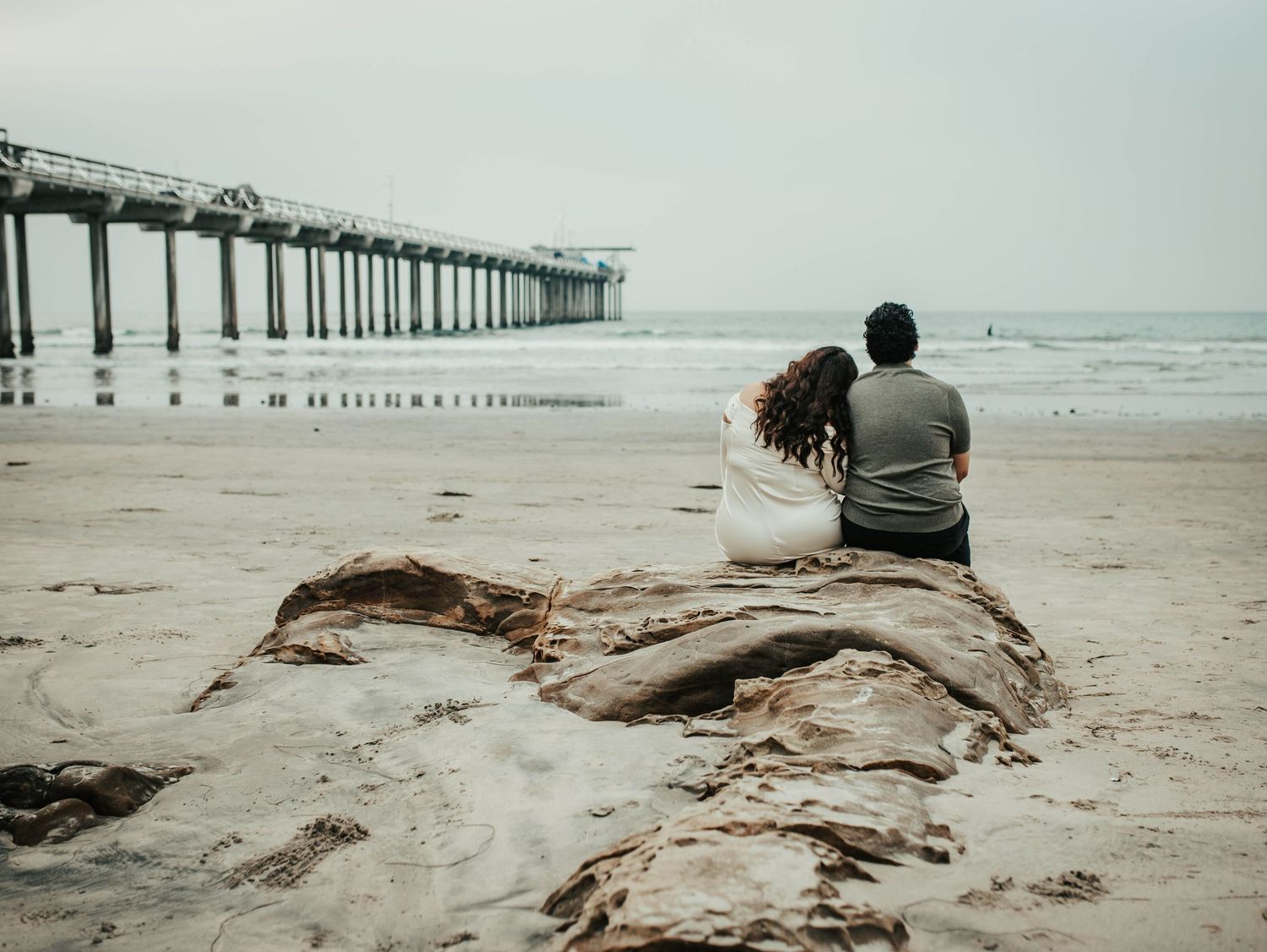 Scripps Pier San Diego CA Beach Engagement Photo Session; couple sitting on rock, embracing, facing the beach with pier in background