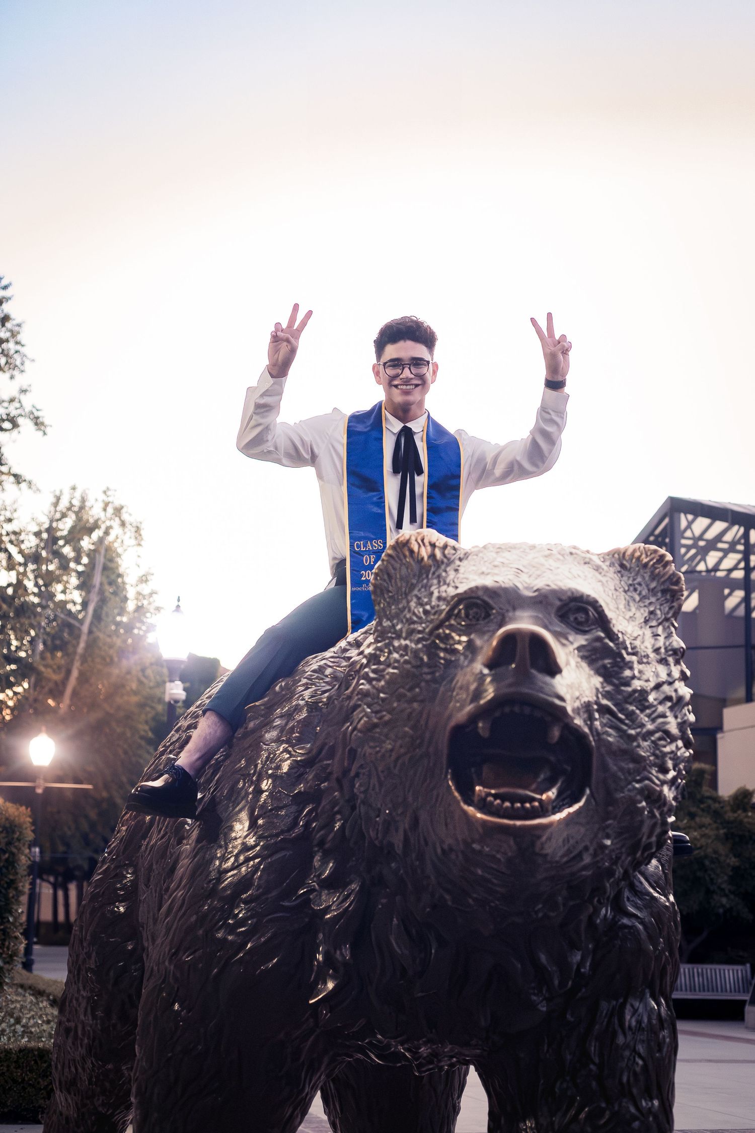 Excited UCLA graduate sitting atop Bruin Bear statue making peace signs at sunset.