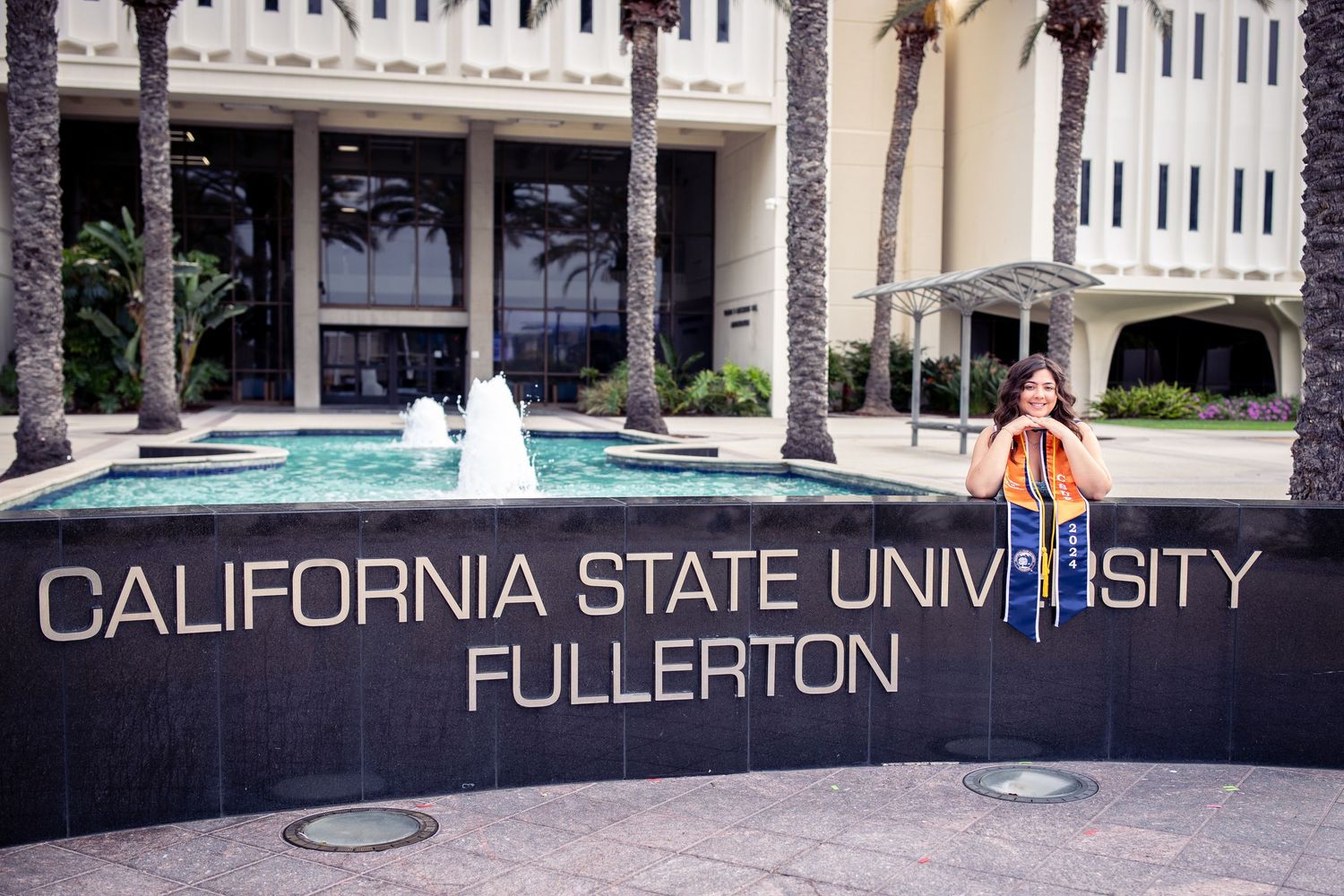 A CSUF graduate, wearing her graduation sash, smiles and leans on a CSUF university sign (on campus) during a graduation portrait session