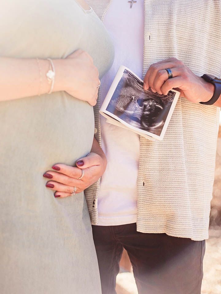 man holds an ultrasound image while the pregnant woman standing next to him is gently embracing her baby bump with both hands