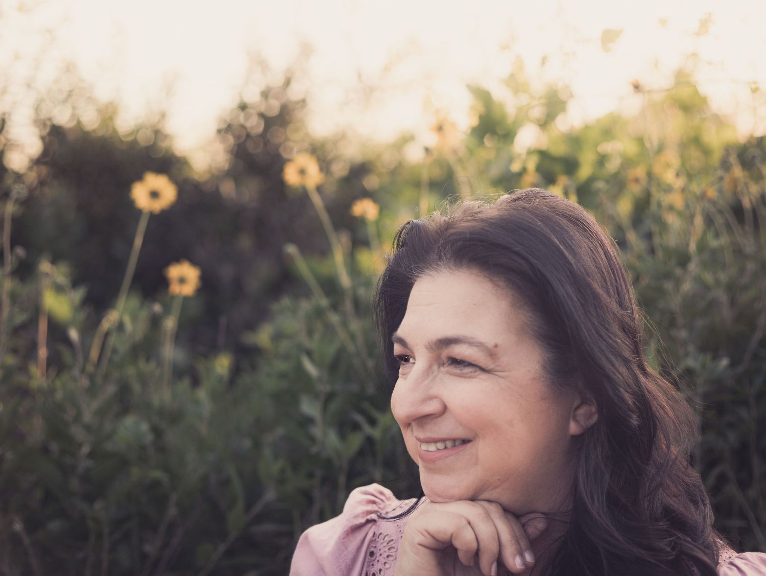 close up of a long-haired woman smiling in a wildflower field of yellow flowers as she looks off-camera, her hand is resting under her chin