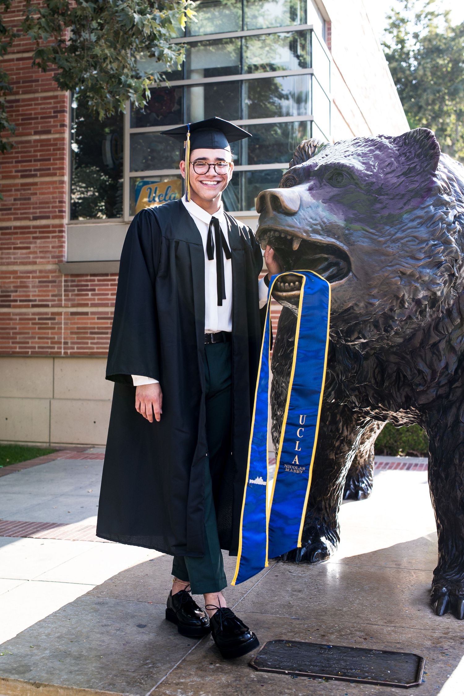 Smiling male UCLA graduate in black gown posing beside the Bruin Bear statue on campus.