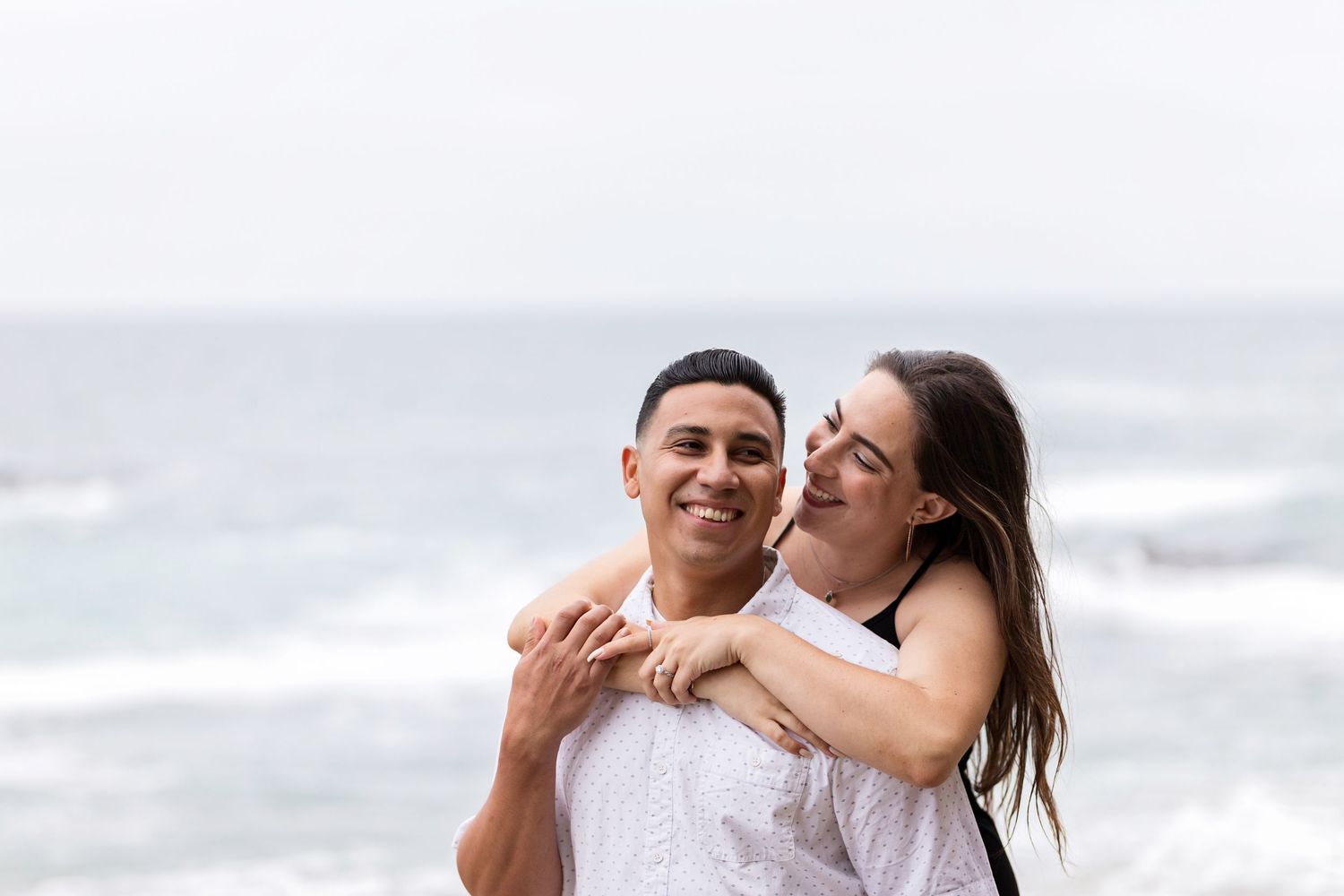 Woman is standing behind a man as she embraces him from behind during a beach engagement session. They are smiling at each other.