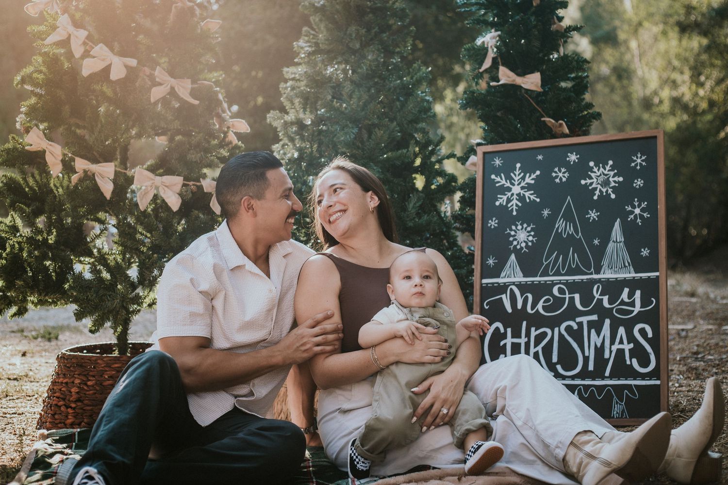 Family of three posing with a Merry Christmas chalkboard sign outdoors among trees for a festive holiday photo.