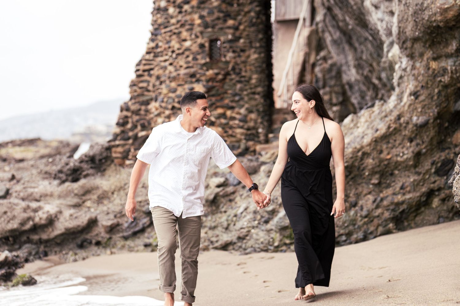 couple holding hands, looking at each other and smiling while walking on the shore during a Pirate's Cove Laguna Beach engagement session