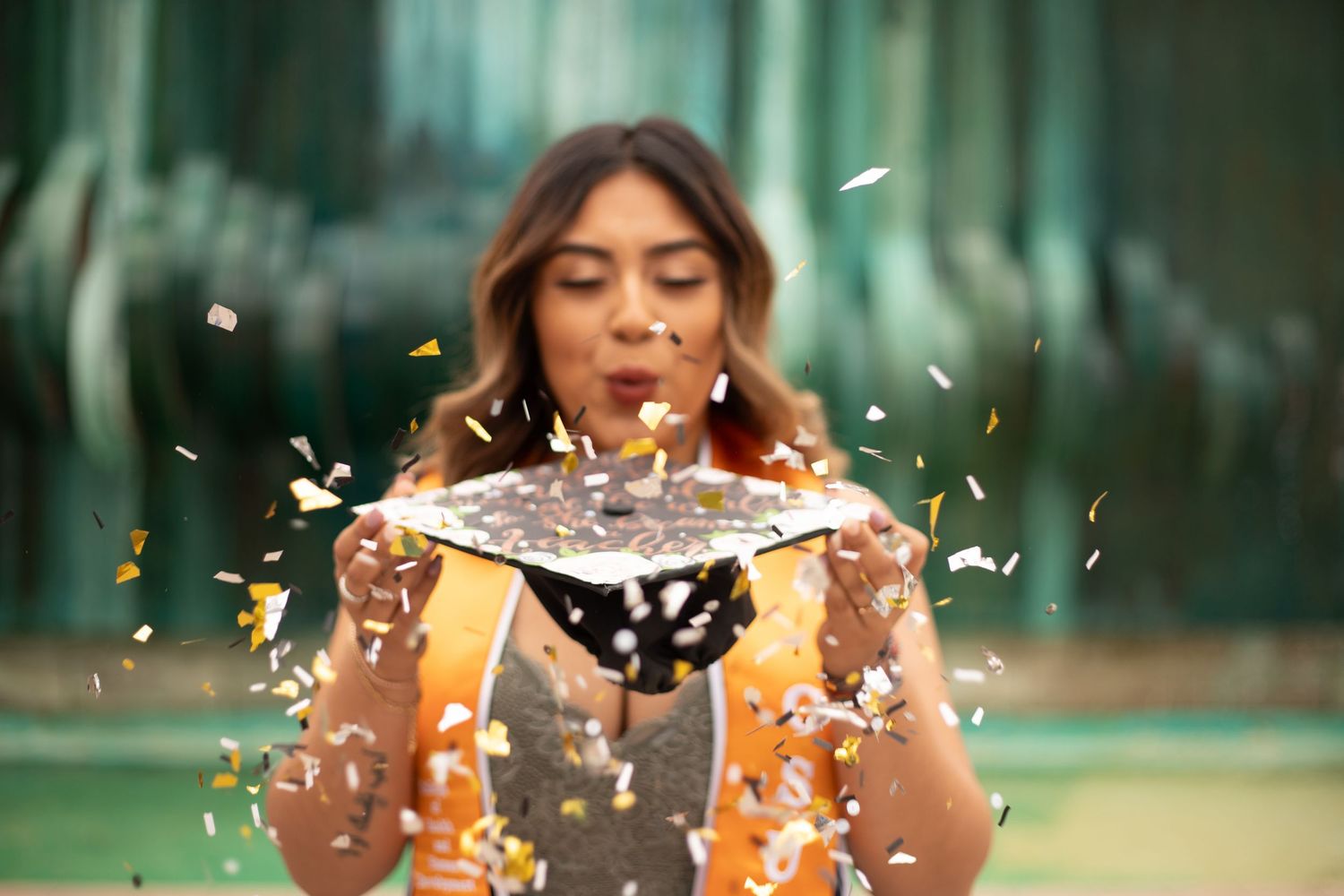 fun graduation photo at CSUF of a graduate holding her cap in front of her as she blows gold glitter off of the cap into the air around her