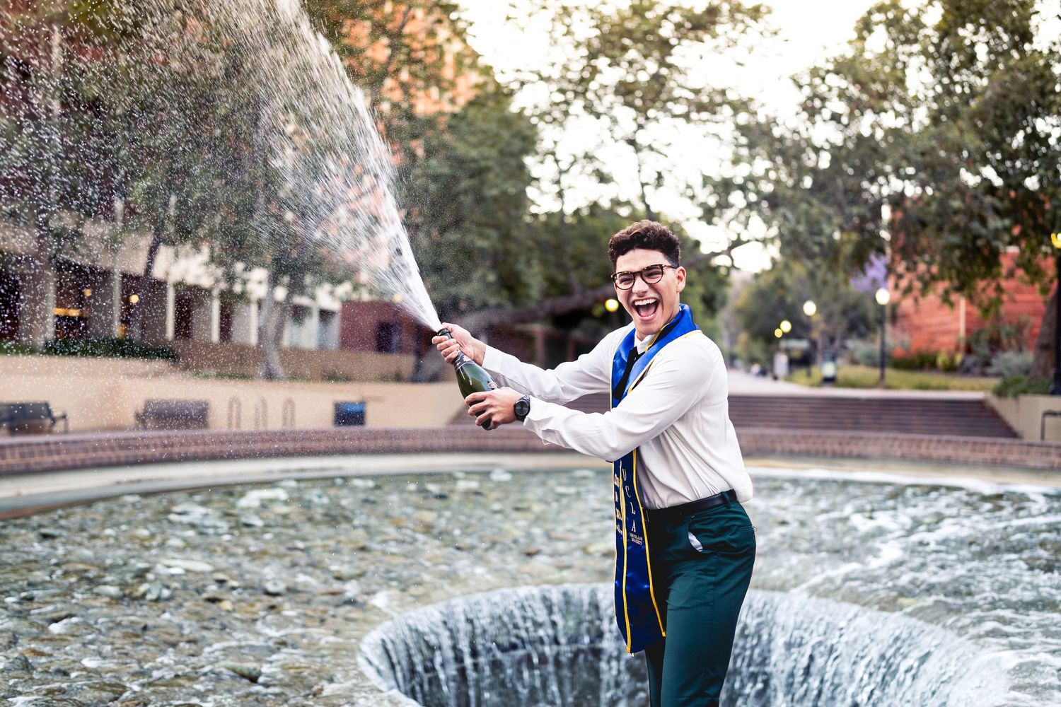 Smiling graduate celebrates by spraying champagne at outdoor campus fountain at sunset.