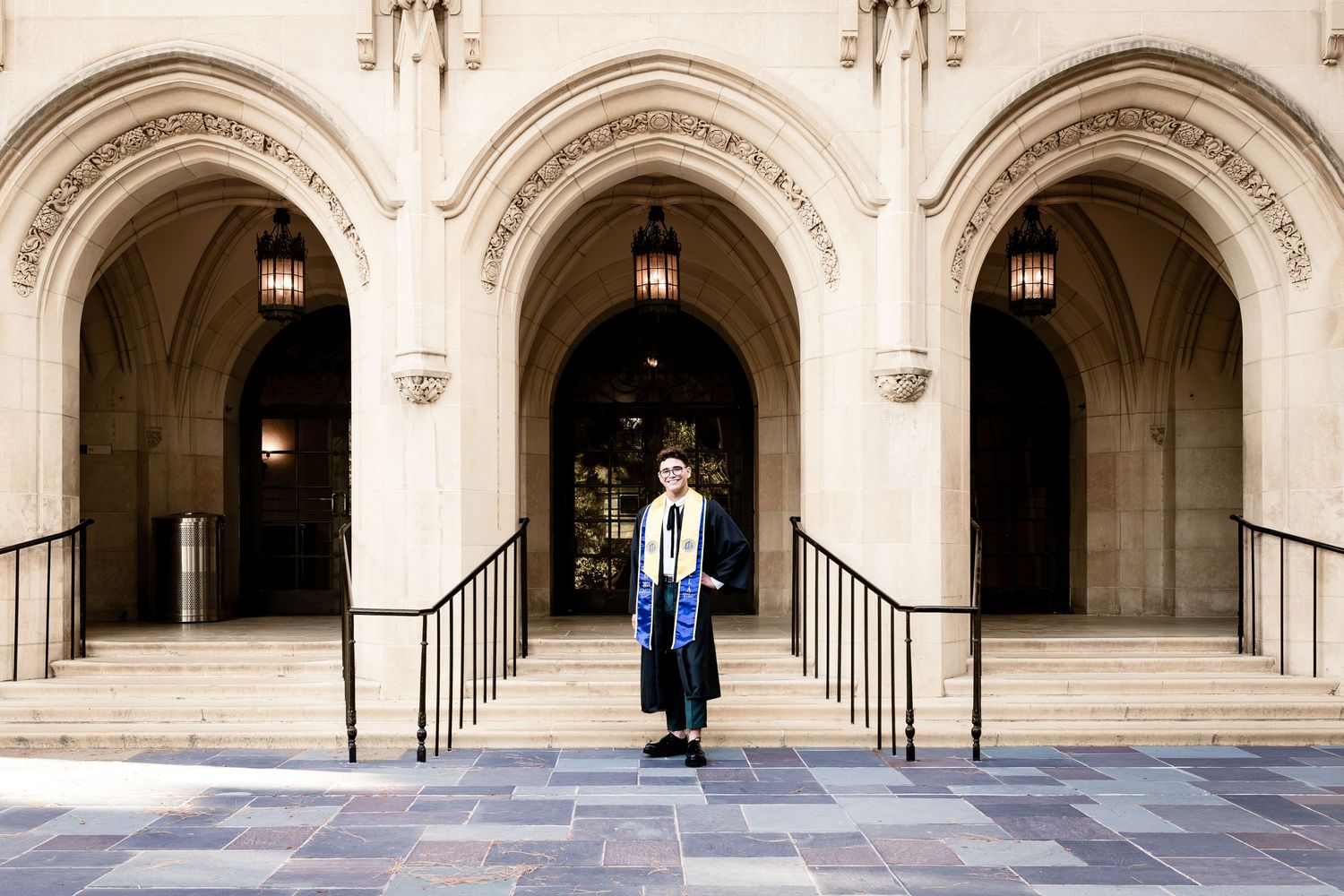 Graduate in cap and gown poses under grand stone archways at university building entrance.