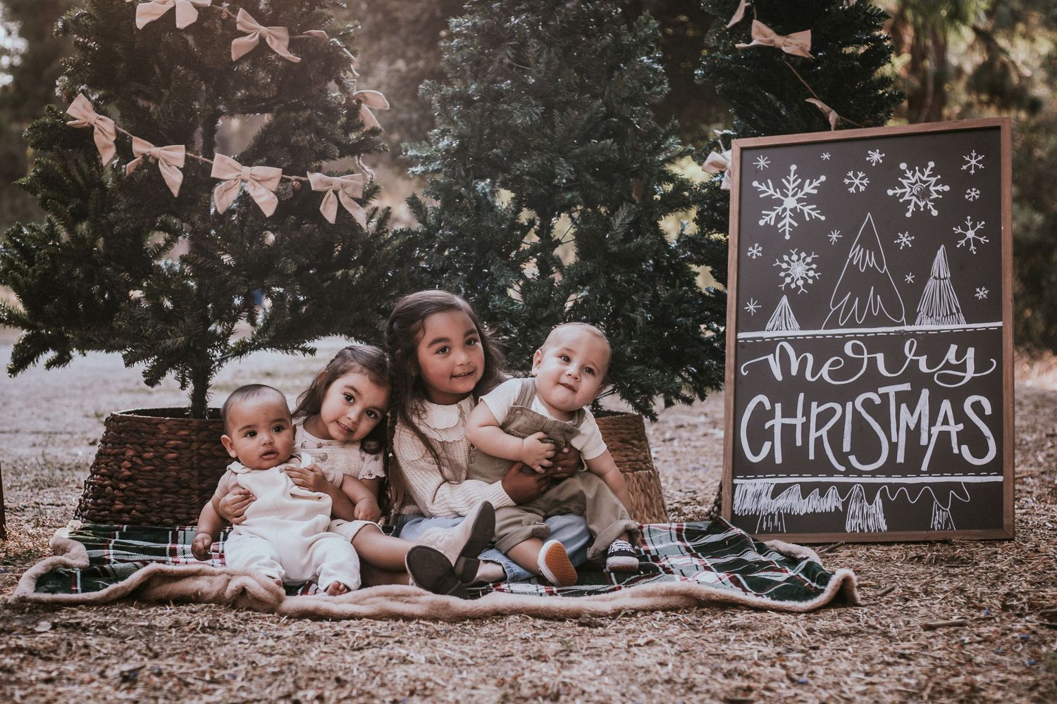Four young children sit together on a blanket outdoors beside a Merry Christmas chalkboard sign surrounded by festive holiday decor.
