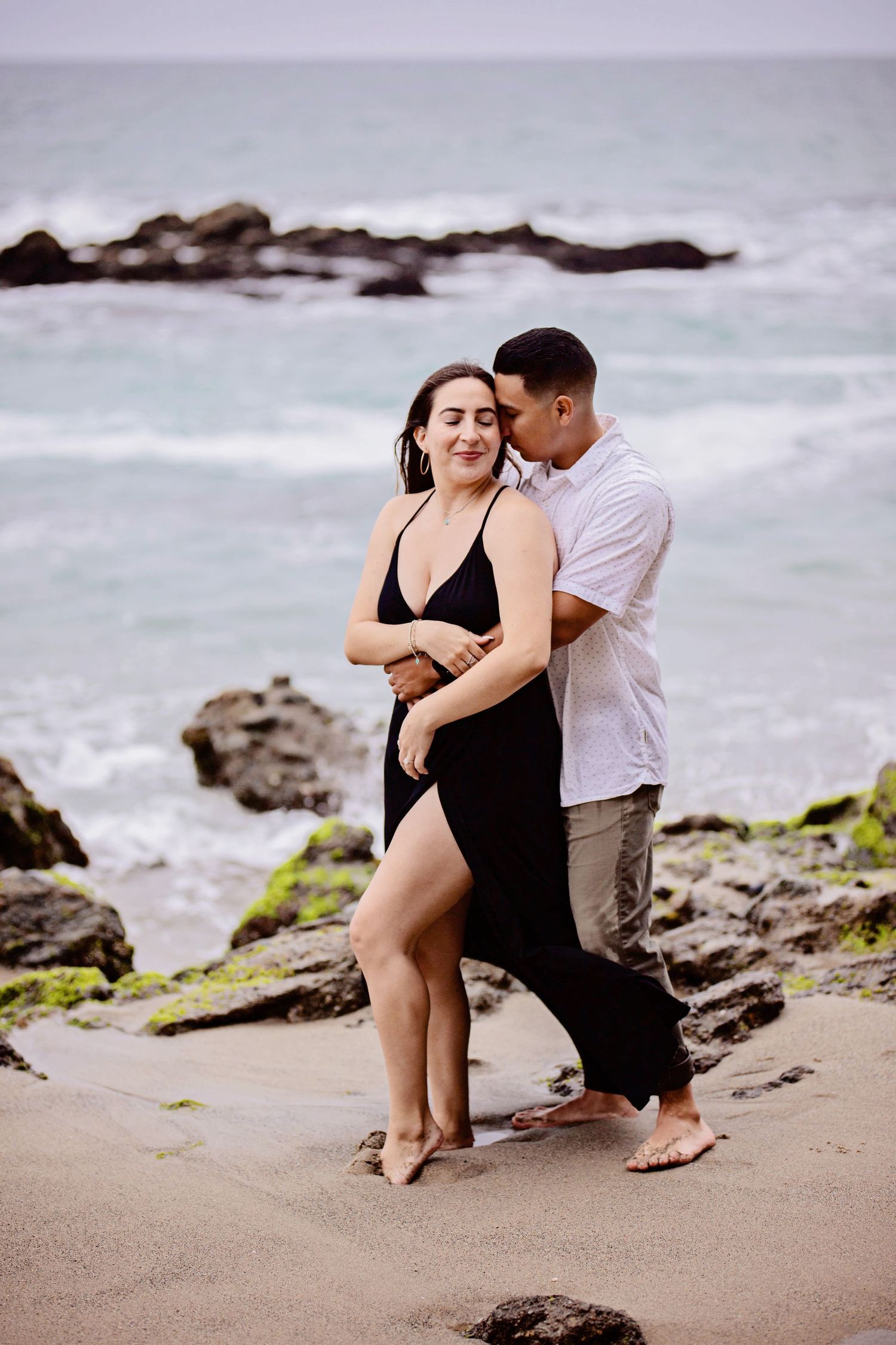 couple tenderly embrace on the sandy shore during a Laguna Beach engagement session. He embraces her from behind as she turns toward him