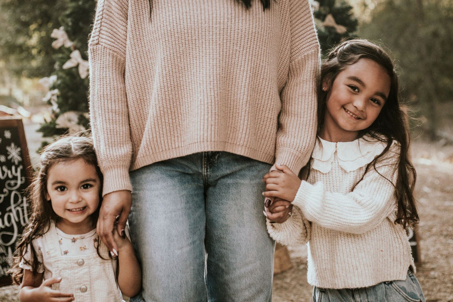 Two young girls smile and hold hands with a woman in a pink sweater and jeans outdoors.