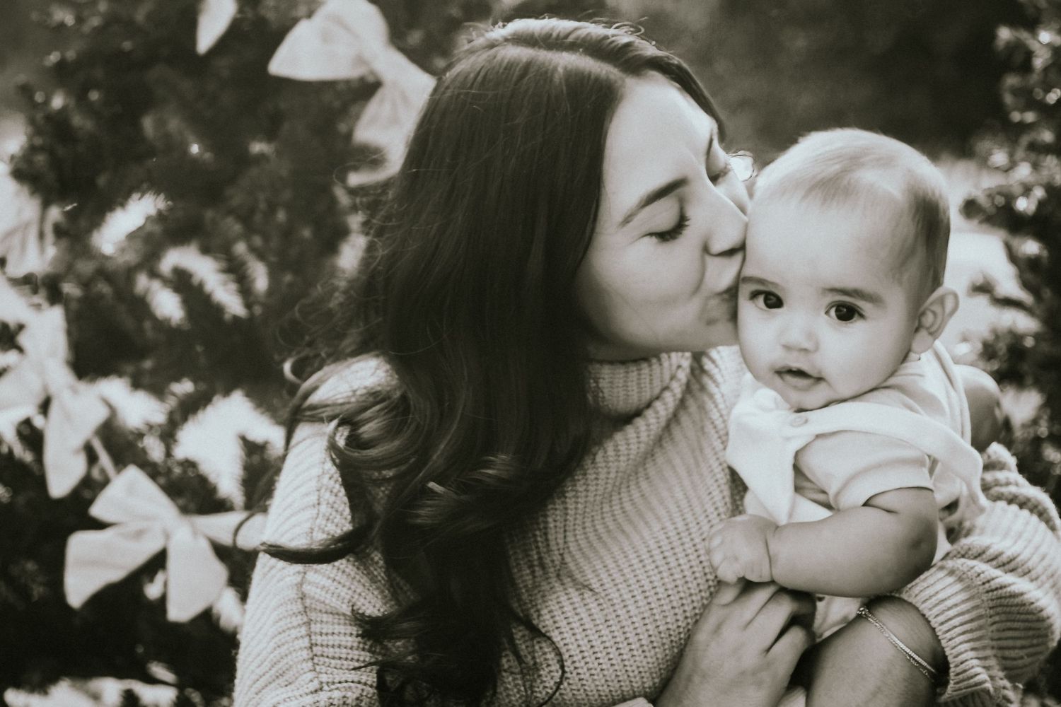 Black and white photo of a young mother kissing her baby on the cheek while holding them outdoors.