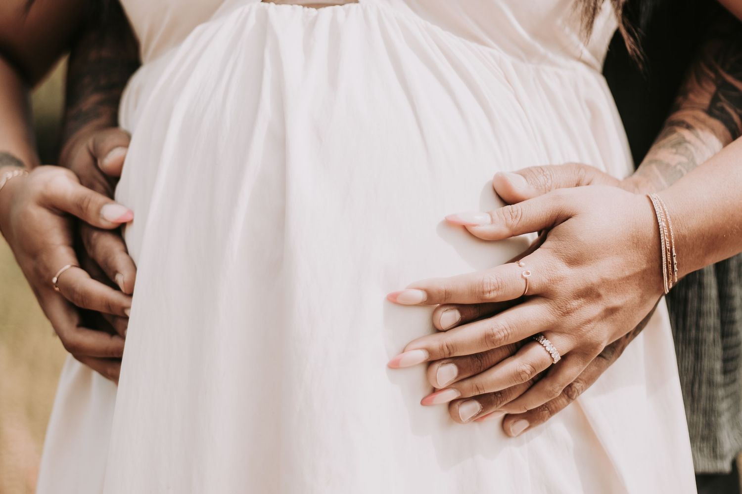 close-up photo of baby bump as pregnant couple holds hands and embraces the baby bump. Woman is wearing a white, flowy dress.