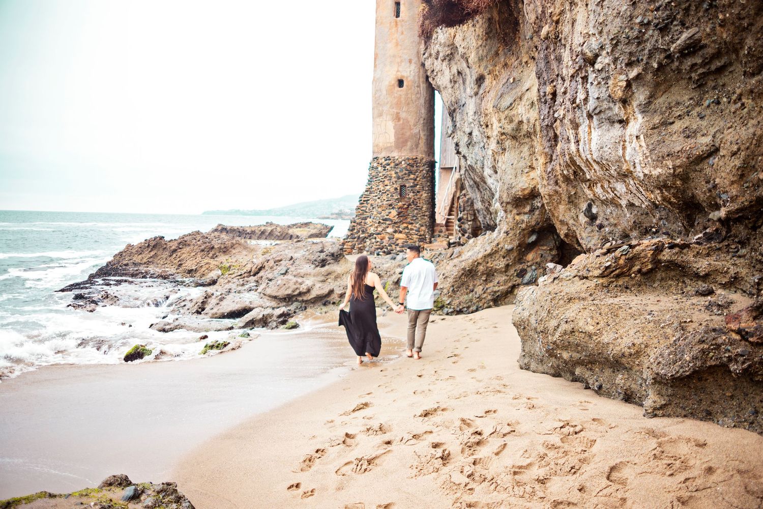 wide shot of a couple holding hands on the beach walking on the shore, backs facing the camera during an engagement session at Laguna Beach