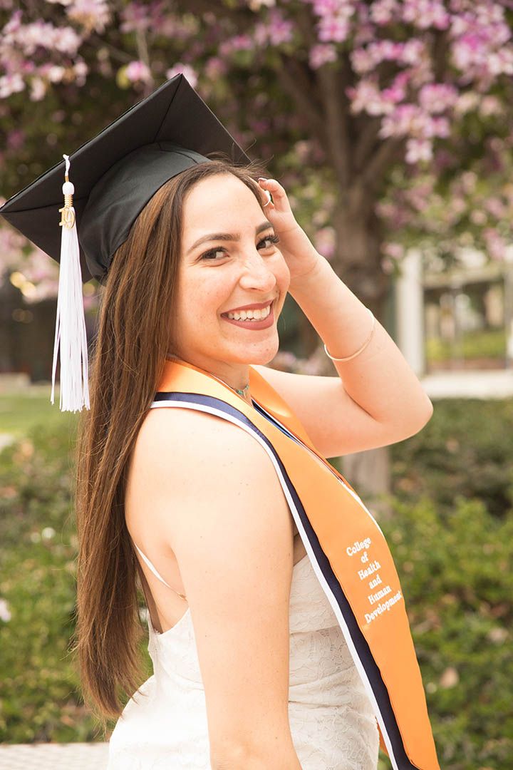 CSUF graduate wearing a white dress, with her graduation cap and tassel on, turning toward the camera and smiling during a graduate session