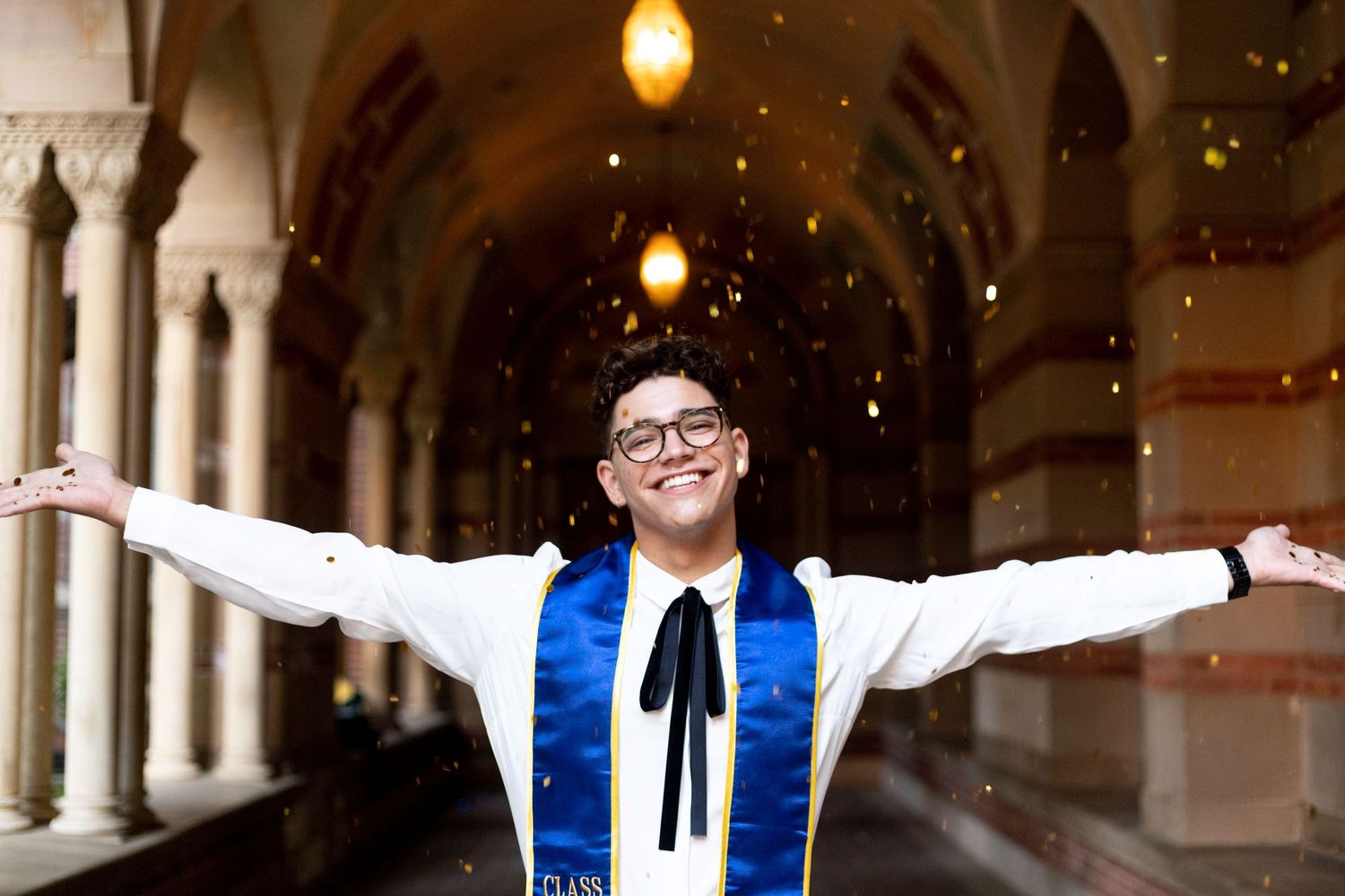 UCLA graduate smiling and standing with arms open wide in an ornate hallway as golden glitter glows all around him in a bokeh effect