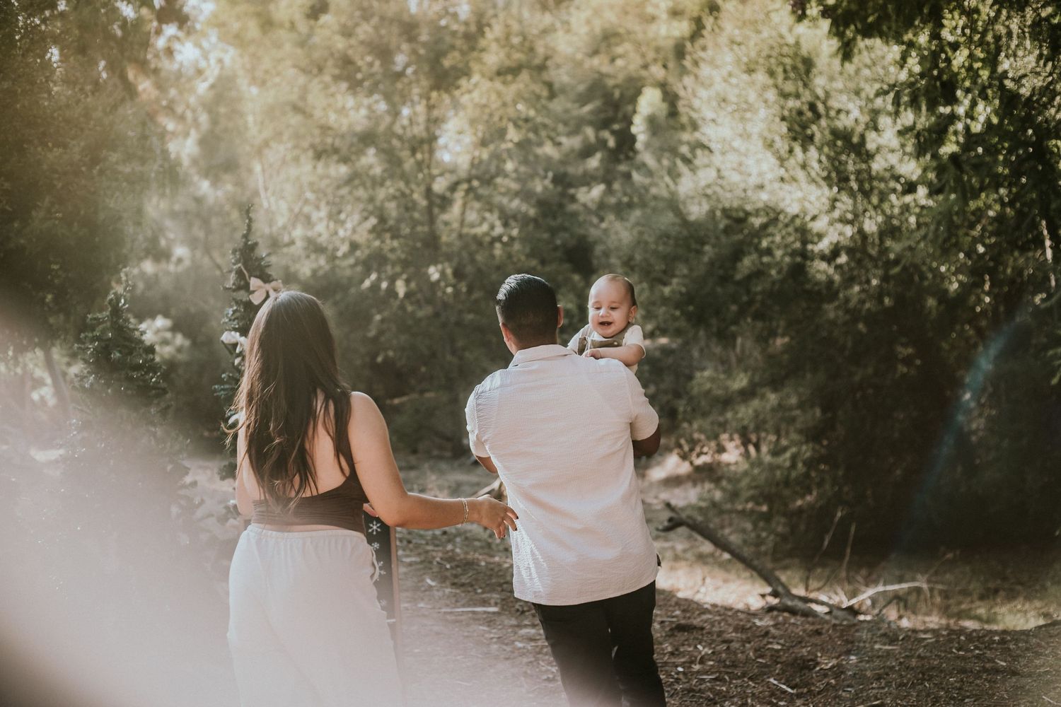 A couple holds their baby outdoors in a sunlit forest setting during a family photo session.