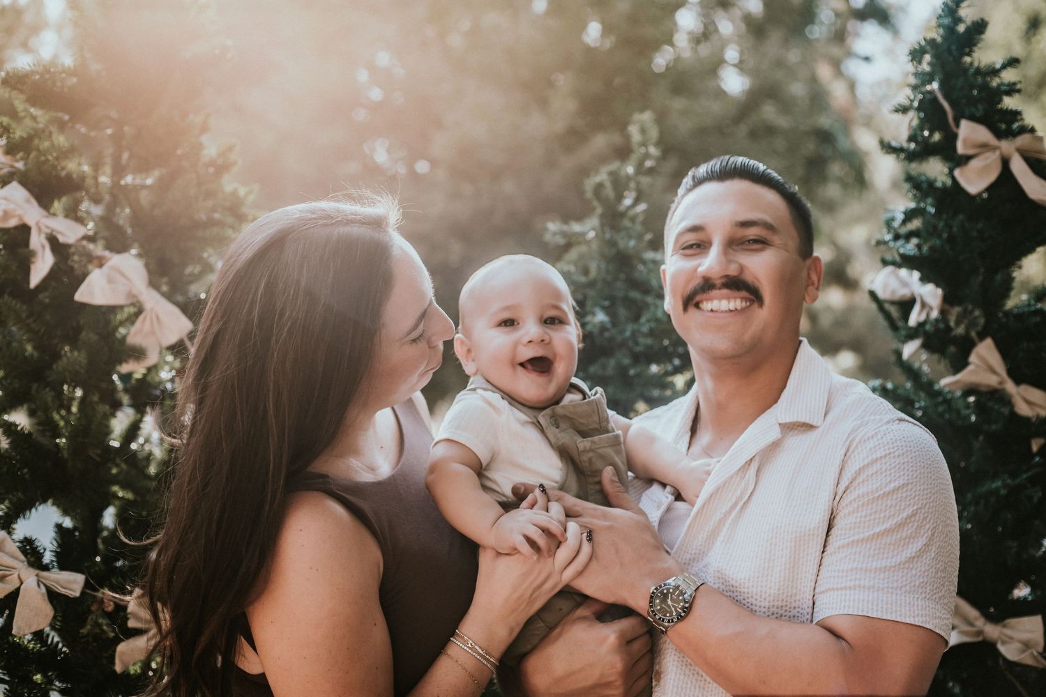 Happy family with smiling baby boy posing outdoors near decorated Christmas trees in warm golden sunlight.