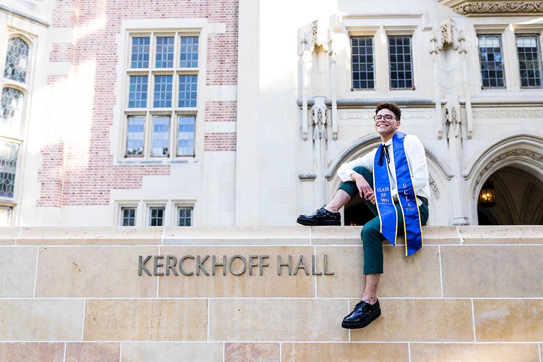 UCLA graduate with his sash, sitting on a ledge in front of Kerckhoff Hall, smiling at the camera