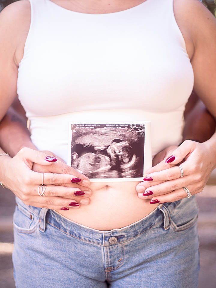 Pregnant woman with crop top stands as partner stands behind her; they hold hands on her baby bump while showing an ultrasound image
