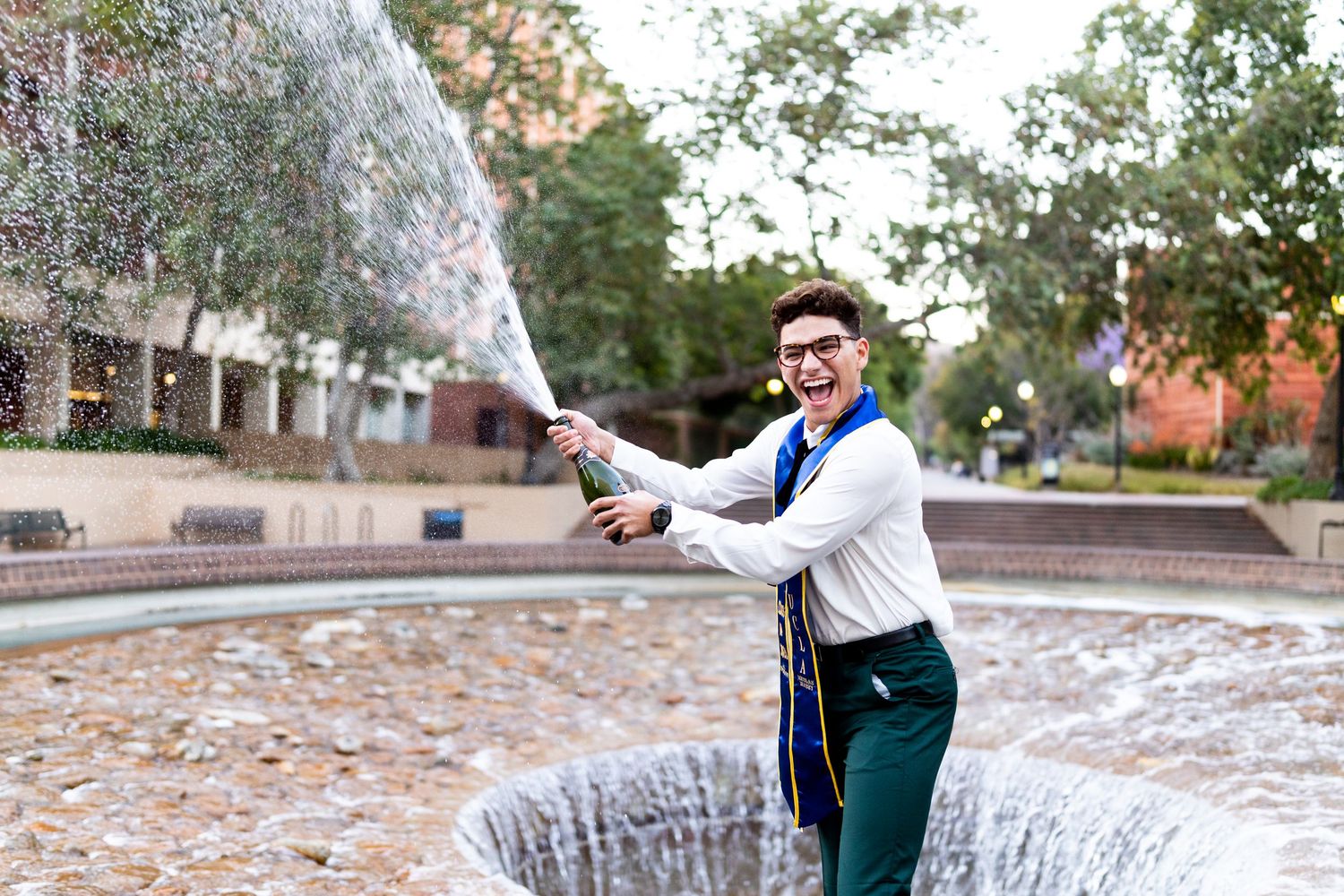 A UCLA graduate standing in the inverted fountain smiles at the camera and laughs as he pops champagne during a graduation portrait session.
