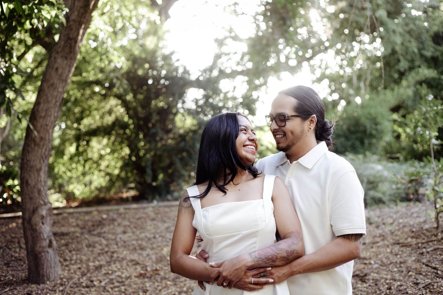 Couple embracing and smiling at each other during an outdoor engagement session in a wooded park with soft light filtering through trees