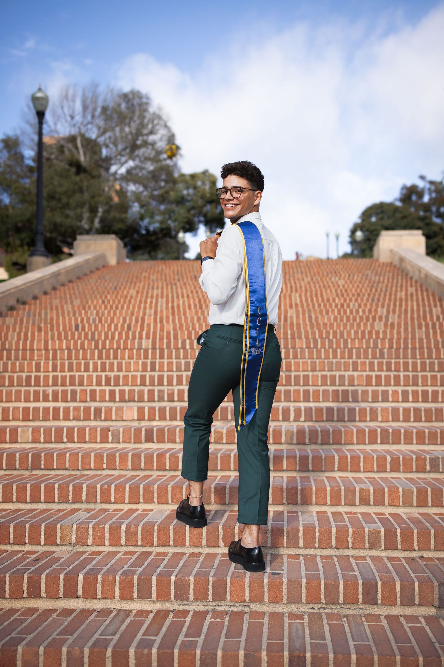 Graduate in green pants and blue stole walks up brick stairs smiling over shoulder.