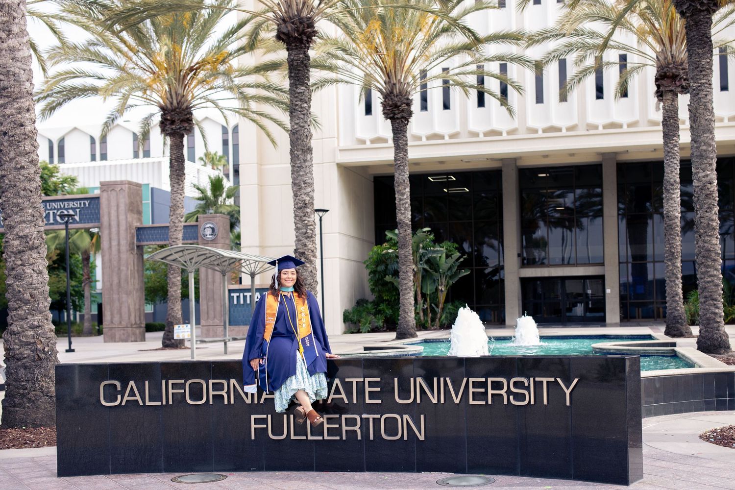 CSUF graduate sits on a CSUF college sign in full graduation attire as she smiles at the camera. Palm trees are in the background.