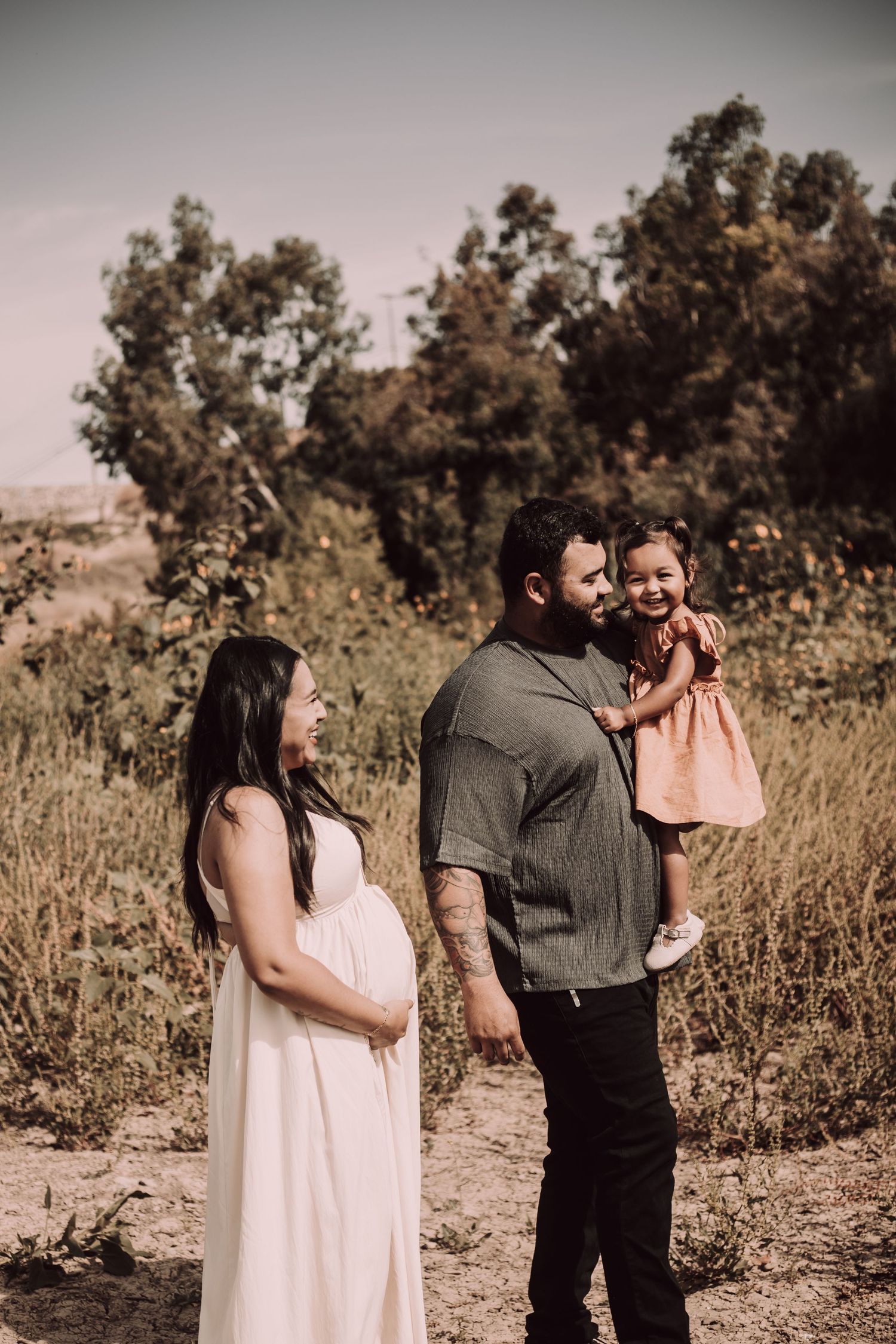 Young expecting couple walks in a rural area. The man is holding their toddler daughter. Mom is looking at and smiling at her daughter