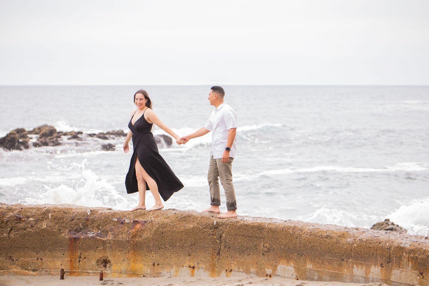 Couple holding hands and smiling while walking along a stone ledge overlooking the ocean during a Laguna Beach engagement session